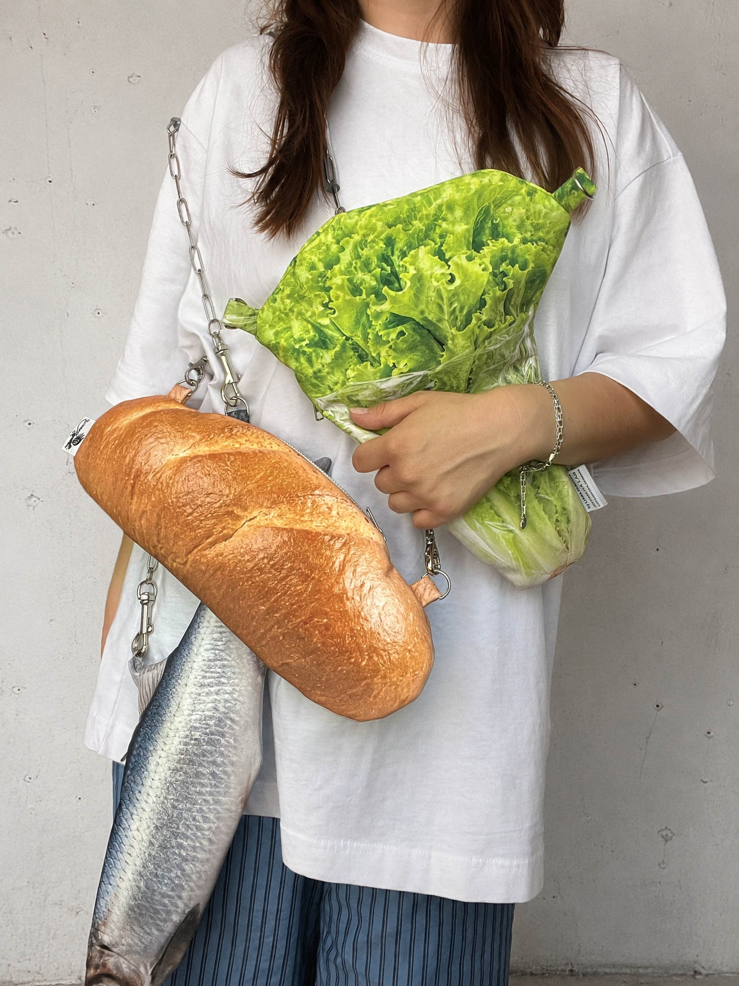 A person wearing an oversized white T-shirt holds a crossbody bag shaped like a loaf of bread, with a fish-shaped bag and a green lettuce-shaped pouch bag hanging from silver chain straps, against a light gray wall.