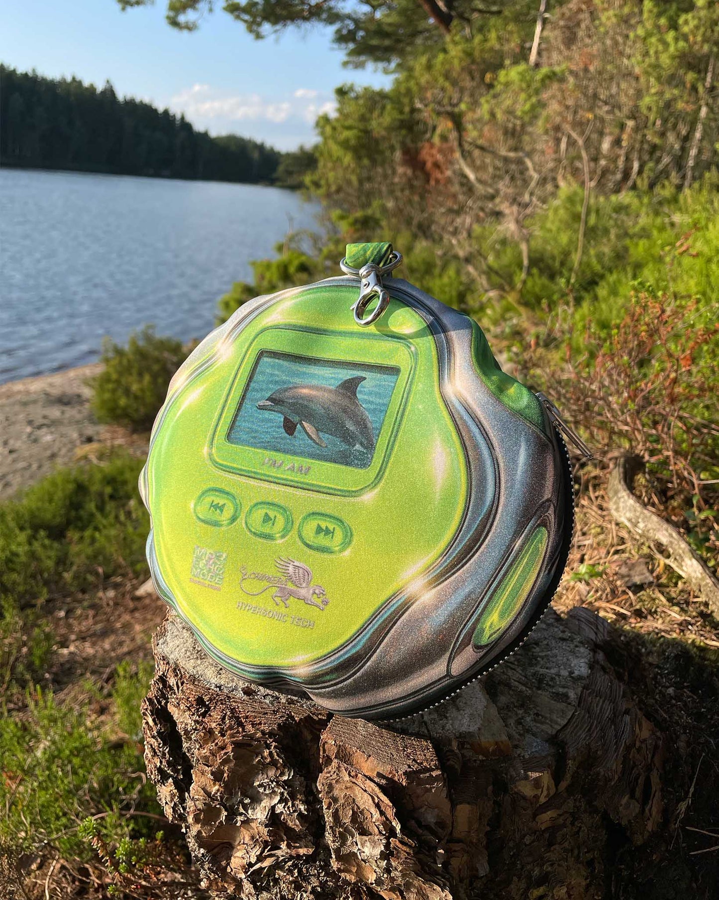 DOLPH MP3 pouch bag by Chimera Lab placed on a tree stump near a lake, showing vibrant green dolphin graphic and retro MP3 player design.