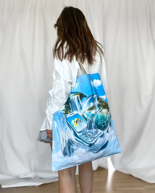 Model wearing the Dreamline OS Tote Bag with Y2K and Frutiger Aero inspired print. Print featues tropical globes with waterfalls and beach scene, surreal phone and yellow fish print, full body shot.