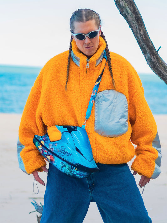 Person wearing a bright orange sherpa jacket and sunglasses, modeling the Chimera Lab x Sharkwork jet ski-shaped bag on a beach.