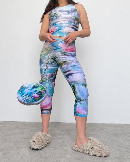 A model wearing a matching two-piece activewear set with a vibrant, multicolored nature print featuring waterfalls, mountains, and pink lotus flowers. She is also holding a matching printed pouch bag and wearing fluffy beige slippers against a plain white wall and gray floor.
