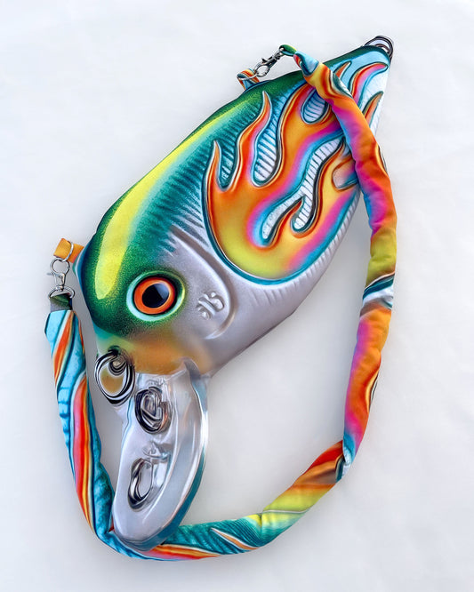 Close-up of the Moto Fish Bag by Chimera Lab, shaped like a 3D fishing lure with a chrome body and neon flame graphic, featuring a padded printed strap.