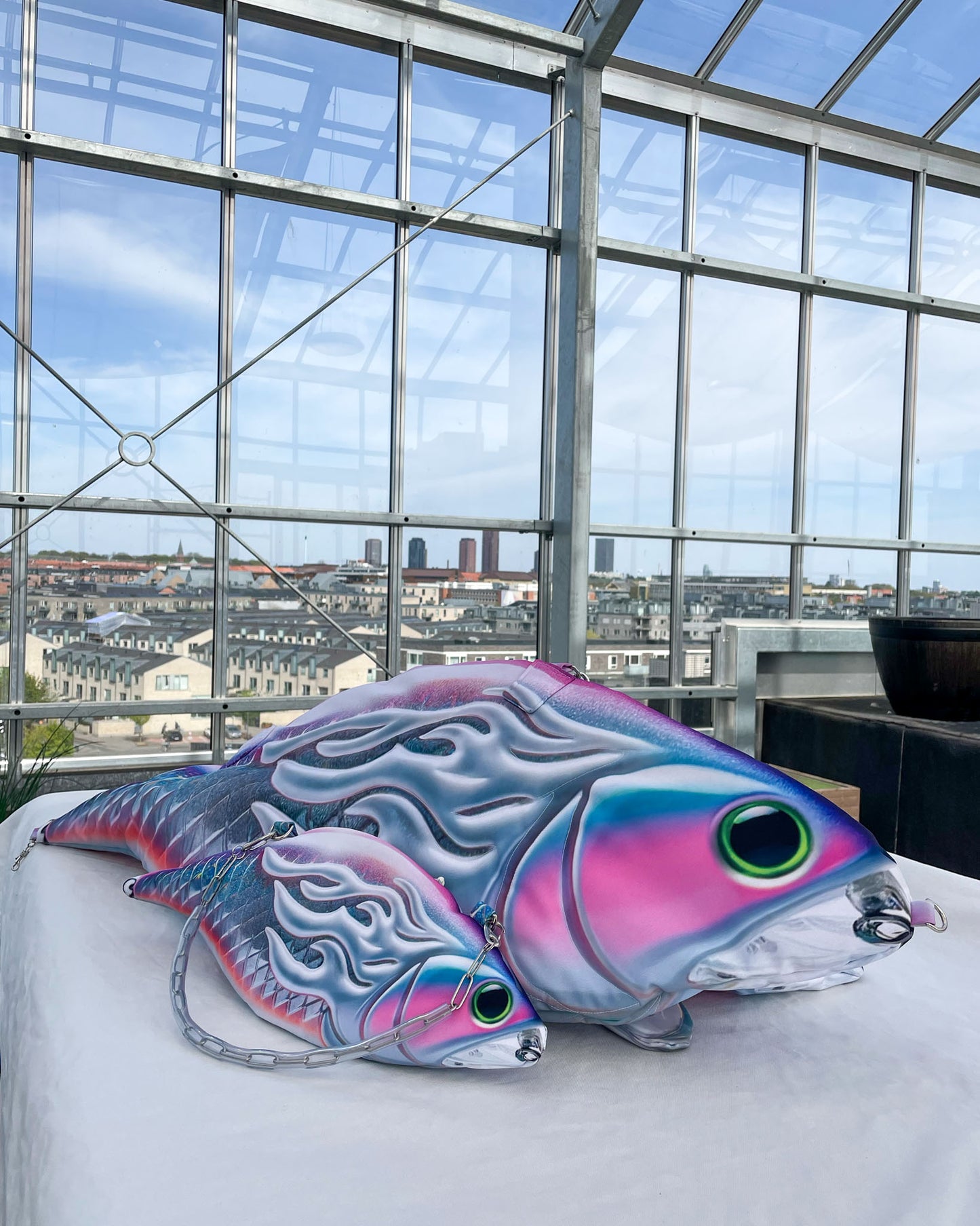 XXL and mini fish-shaped handbags with iridescent 3D-effect prints and metallic flame detailing, displayed together on a rooftop with a cityscape view in the background. Each bag features bold pink, silver, and purple tones with neon green eyes and detachable straps.
