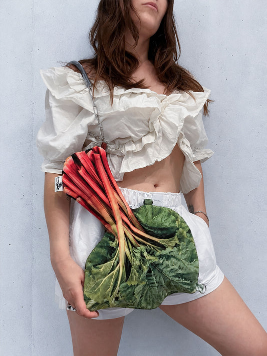 A model wearing a white ruffled crop top and white shorts holds a green leafy bag that resembles a bunch of fresh rhubarb with red and orange stalks, against a light gray wall.