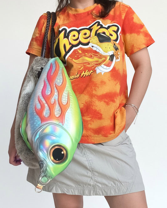 Model wearing Cheetos graphic tee and khaki skirt, carrying the large Trophy Fish Bag with holographic flame print and surreal fish-eye detail.