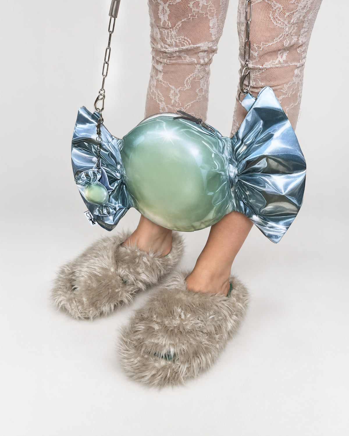 A person wearing fuzzy taupe slippers and sheer lace-patterned tights holds a metallic candy-shaped purse. The purse has a large iridescent green center wrapped in shiny silver-blue foil-like printed material with silver chain straps, photographed against a clean white studio background.