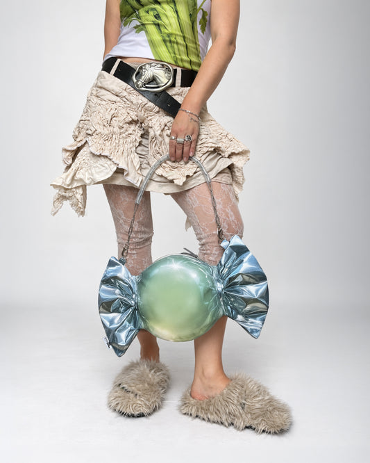 A person wearing a green and white top, a beige ruffled skirt, sheer tights, and fuzzy slippers holds a unique candy-shaped handbag with a metallic silver bow and a glossy light green center print, against a plain light gray studio background.