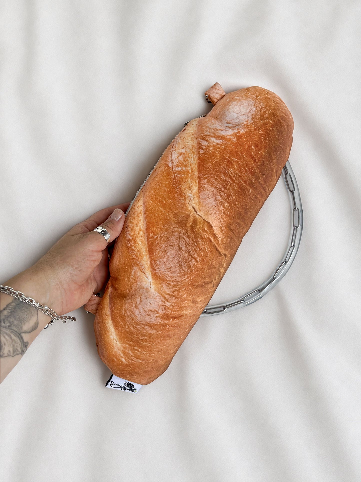 A novelty clutch purse shaped like a baguette of bread, held by a hand against a white fabric background.