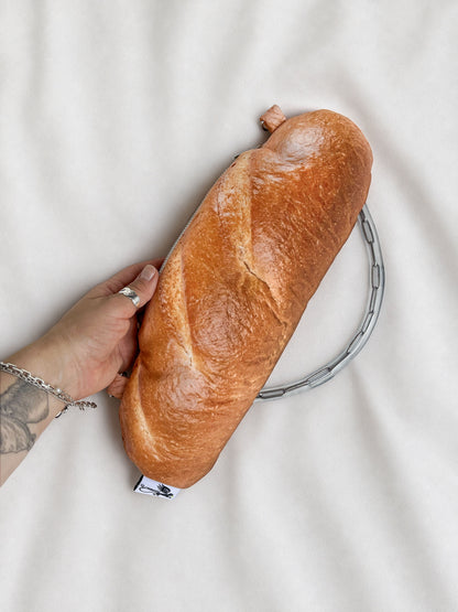 A novelty clutch purse shaped like a baguette of bread, held by a hand against a white fabric background.