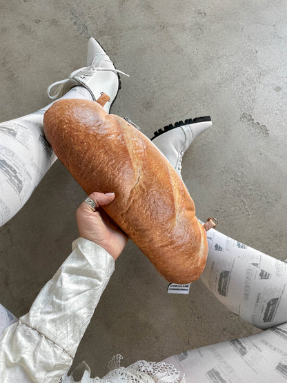 A person holding a large, realistic-looking golden-brown novelty bread loaf bag, wearing white pointed-toe lace-up shoes and white pants with black print, against a concrete floor.