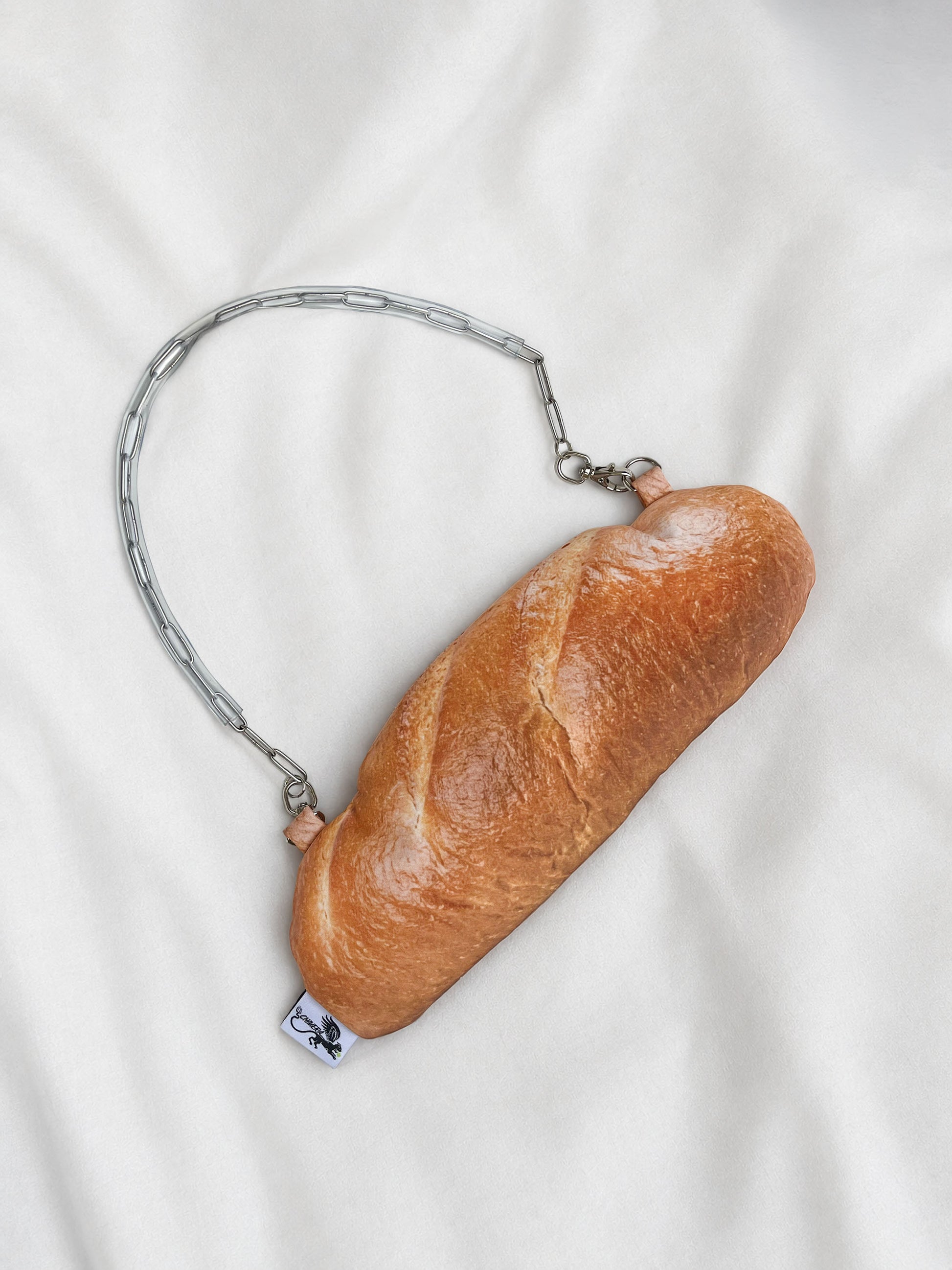 A novelty bread baguette-shaped clutch purse with a silver chain handle, resting on a white fabric background.