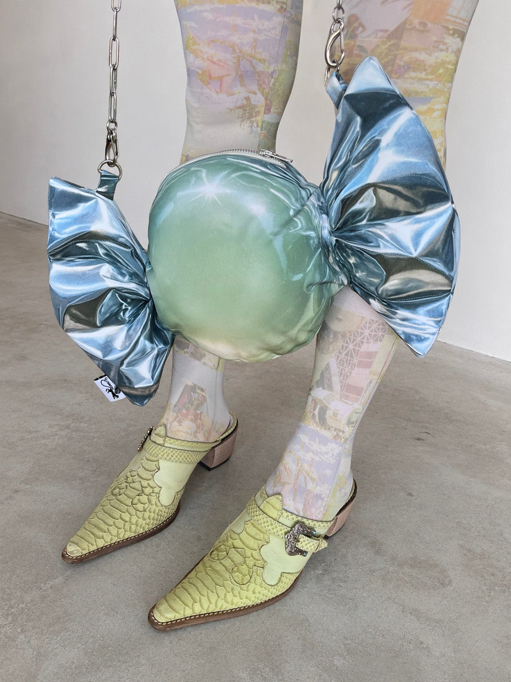 Person wearing a candy-shaped bag in a silver metallic wrap print and pointed shoes on a plain background