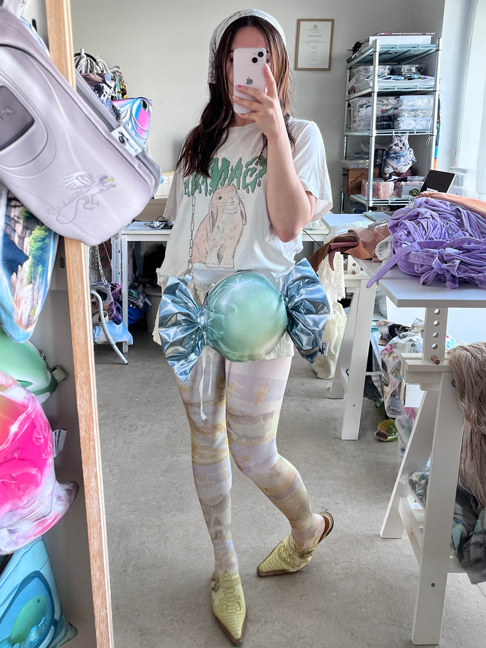 Person wearing a white graphic t-shirt and colorful leggings, styled with an oversized wrapped candy print bag, in a fashion studio workroom.