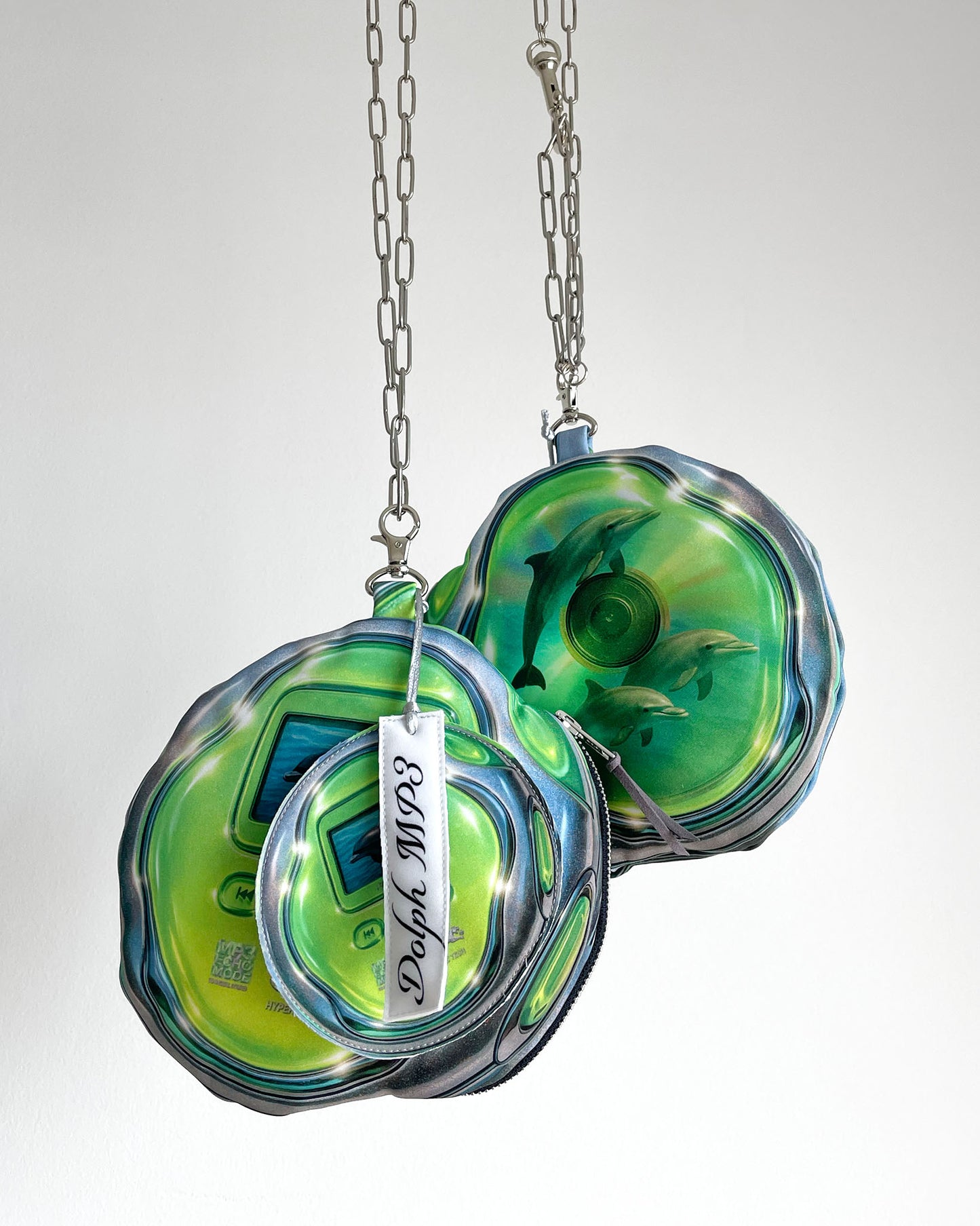 Two DOLPH MP3 pouch bags by Chimera Lab hanging on stainless steel chains, shaped like retro green MP3 players featuring a translucent CD print with dolphins swimming inside on the back side, surreal Y2K tech-inspired design.
