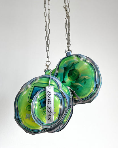Two DOLPH MP3 pouch bags by Chimera Lab hanging on stainless steel chains, shaped like retro green MP3 players featuring a translucent CD print with dolphins swimming inside on the back side, surreal Y2K tech-inspired design.