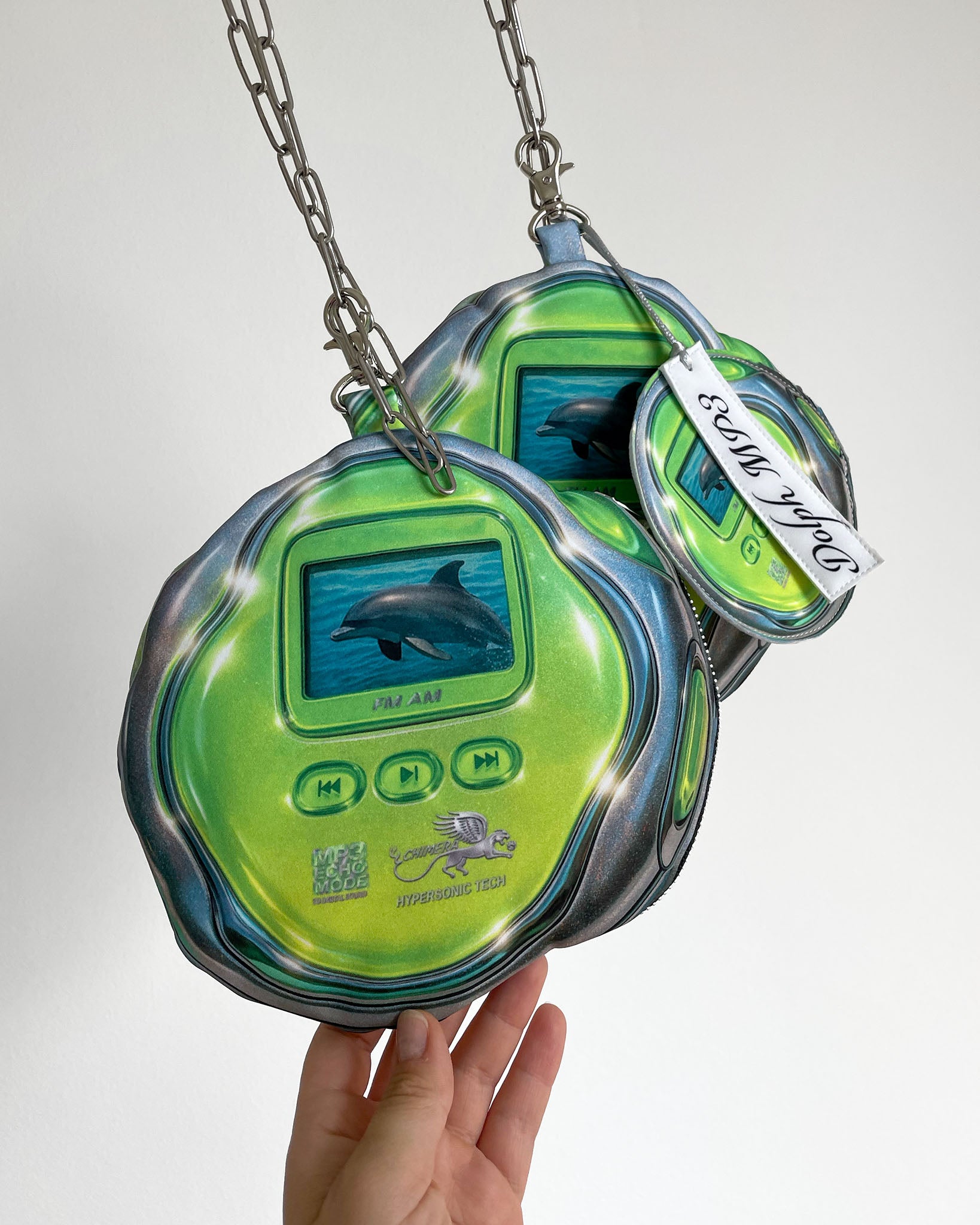 Two DOLPH MP3 pouch bags by Chimera Lab hanging on stainless steel chains, shaped like retro green MP3 players with dolphin graphics, Y2K-inspired digital print accessory.