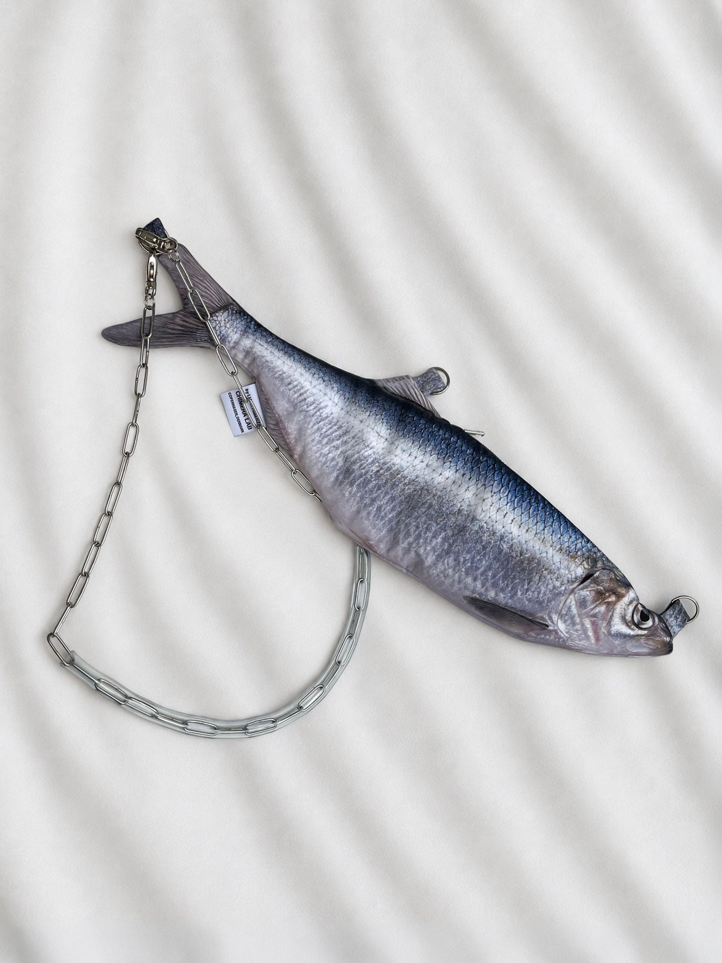 A fish-shaped clutch bag with a silver chain strap, displayed on a white textured fabric background.