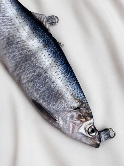 A realistic silver fish-shaped bag with a metal ring at the tail, photographed on a light fabric background.