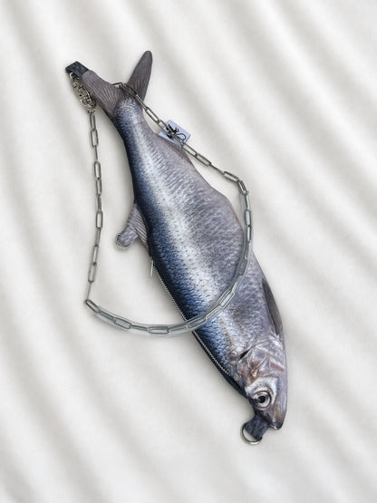 A realistic fish-shaped handbag with a silver chain draped over it, shown on a white textured fabric background.