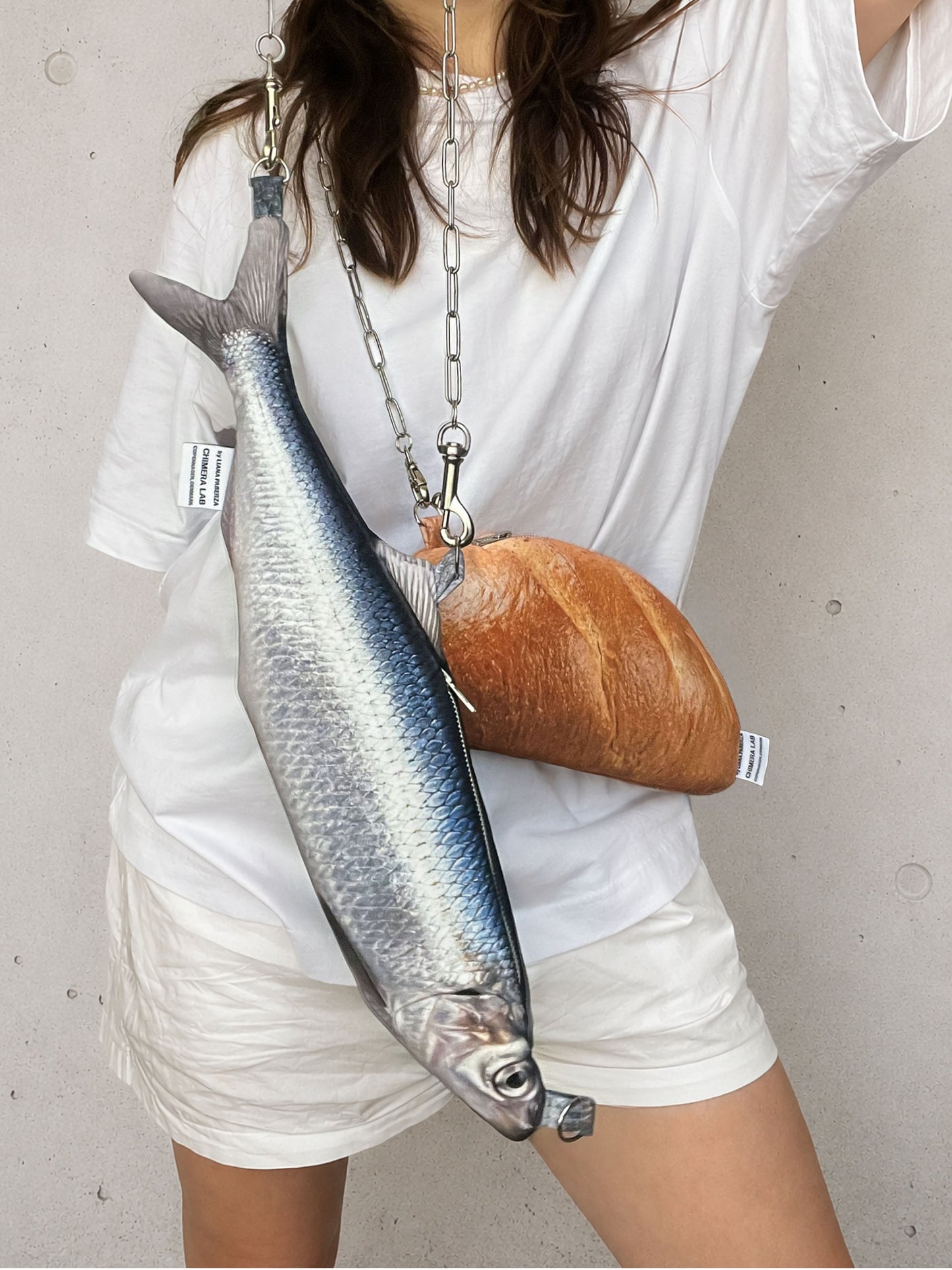 A person wearing a white t-shirt and shorts carries a novelty purse shaped like a whole fish, with a second purse shaped like a loaf of bread hanging from a silver chain across their chest.