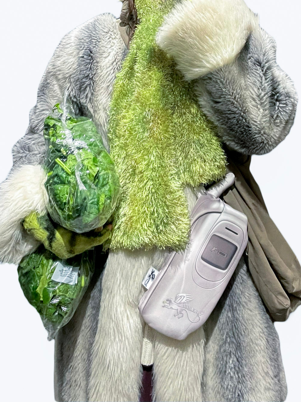 A person wearing a gray faux fur coat with a green scarf, green vegetables, and a small crossbody bag with a silver Y2K flip phone design on a white background.