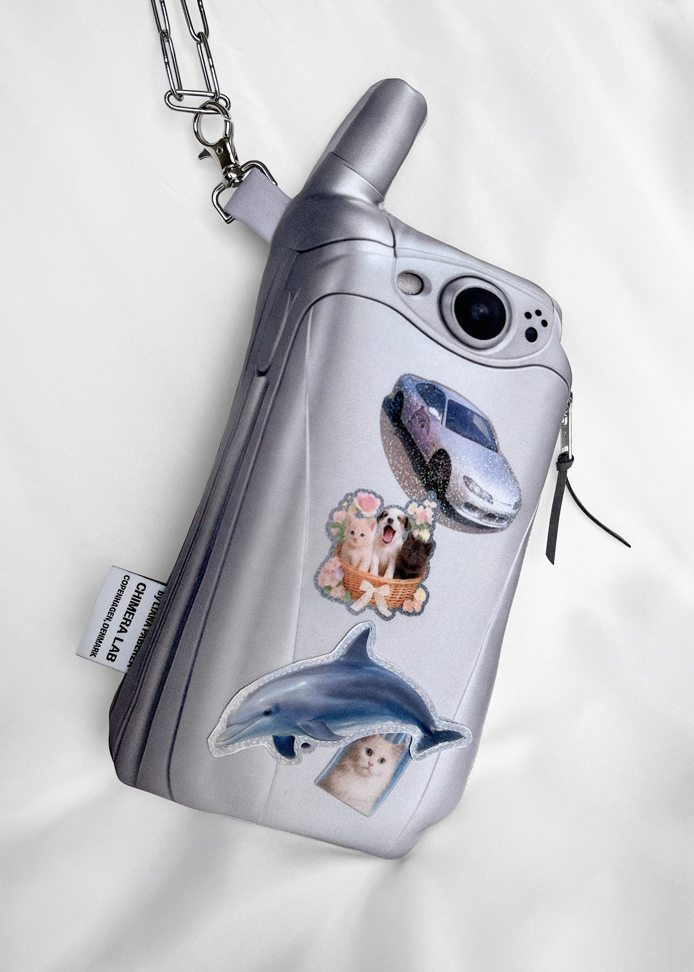 A silver flip phone-shaped pouch with a chain strap, decorated with printed stickers of a car, a dog, a cat, and a dolphin, shown on a white fabric background.