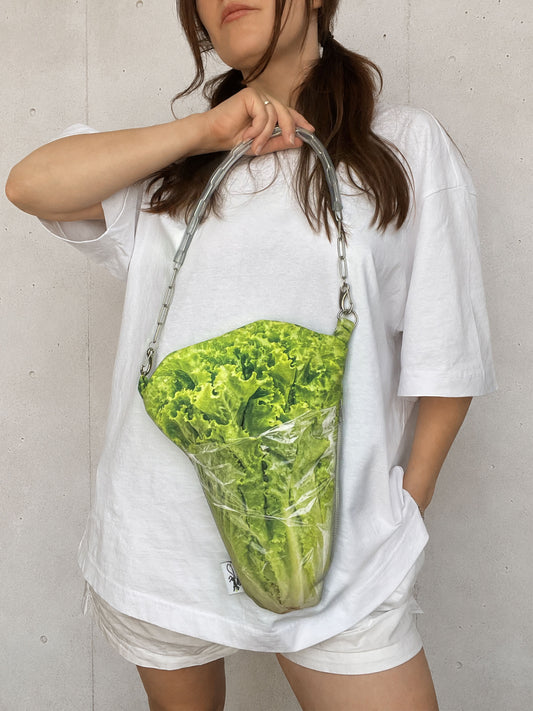 A person wearing an oversized white t-shirt and white shorts holds a crossbody bag shaped like a fresh green lettuce head wrapped in clear plastic, with a silver chain strap, against a light gray concrete wall.