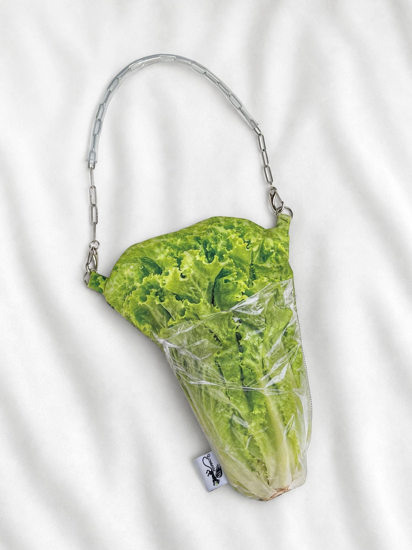 A novelty shoulder bag shaped like a head of lettuce wrapped in clear plastic, with a silver chain strap, laid flat on a white fabric background.