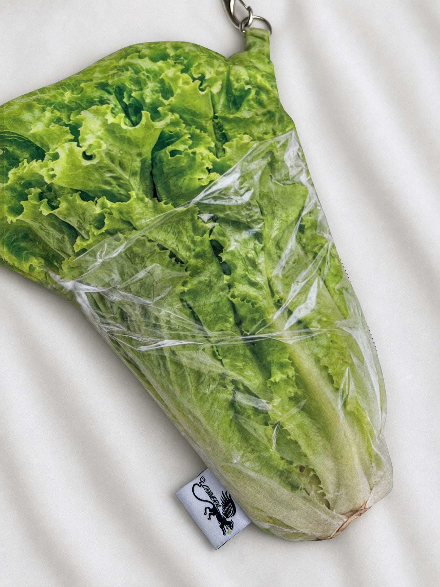 A novelty zipper pouch shaped like a head of lettuce wrapped in clear plastic, shown on a white fabric background. The pouch has a small white brand tag at the bottom with a black logo and a metal ring at the top.