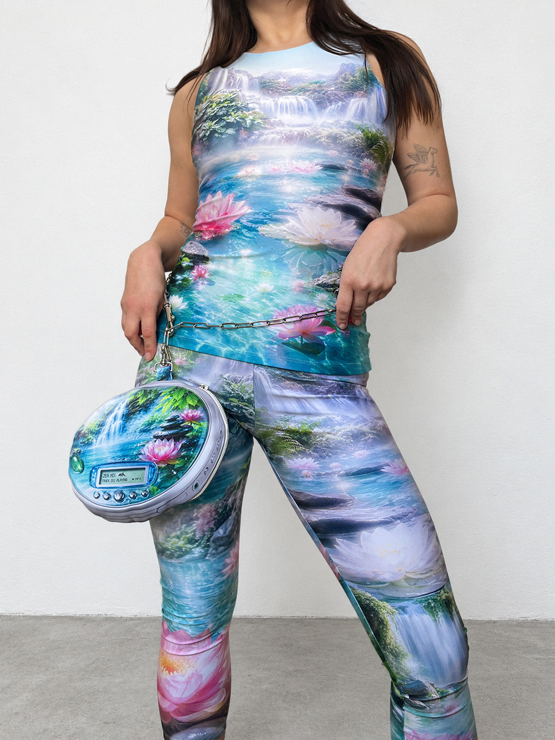 A woman modeling a matching sleeveless tank top and leggings set with a vibrant, multicolored nature print featuring waterfalls, lotus flowers, and ponds. She is holding a matching round CD player-shaped pouch bag.