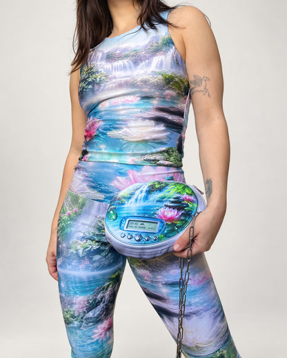 A model wearing a matching sleeveless tank top and leggings set with a vibrant, multicolored nature print of waterfalls, lotus flowers, and ponds. She is holding a matching round shoulder bag with a chain strap.
