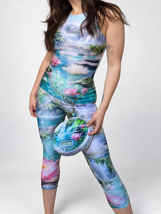 A woman modeling a sleeveless, high-neck, body-hugging tank top and capri-length leggings with a vibrant, full-bleed landscape print of waterfalls, lotus flowers, and ponds. She is holding a small, matching round CD player-shaped pouch bag with stainless steel chain.