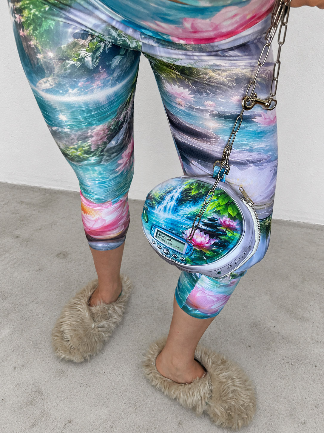 A person wearing colorful zen spa and landscape print leggings, holding a matching round pouch bag, and wearing fluffy beige slippers on a concrete floor.