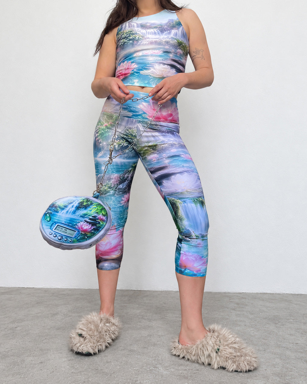 A model wearing a matching two-piece activewear set with a vibrant, multicolored nature print featuring waterfalls, mountains, and pink lotus flowers. She is also holding a matching printed pouch bag and wearing fluffy beige slippers against a plain white wall and gray floor.