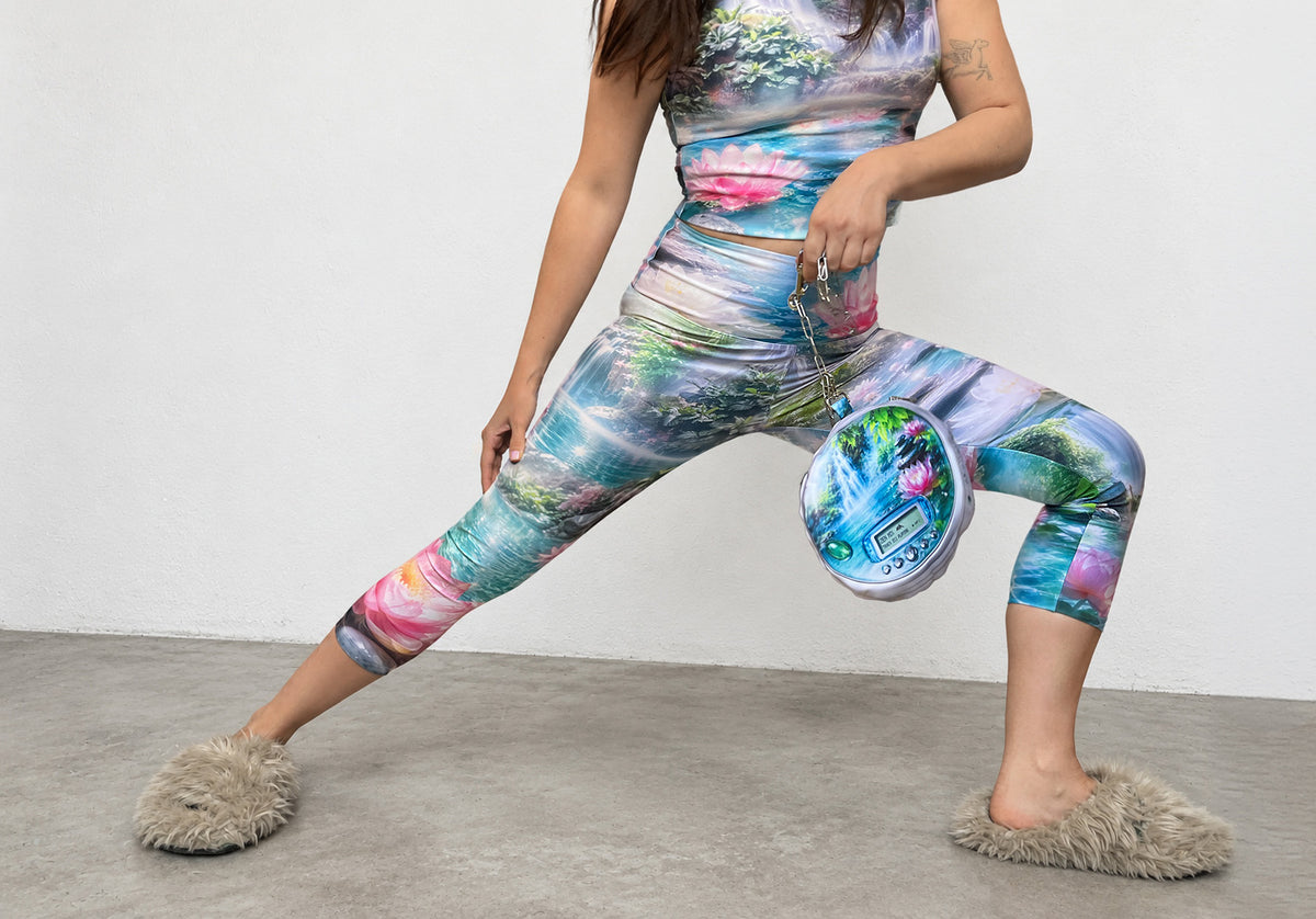 A model wearing a matching two-piece activewear set with a colorful landscape print, posing in a wide stance on a gray concrete floor against a white wall.