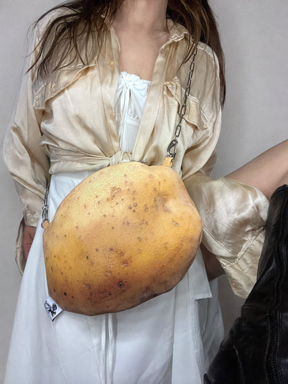 A person wearing a cream satin shirt and white dress carries a large, potato-shaped shoulder bag with a chain strap.