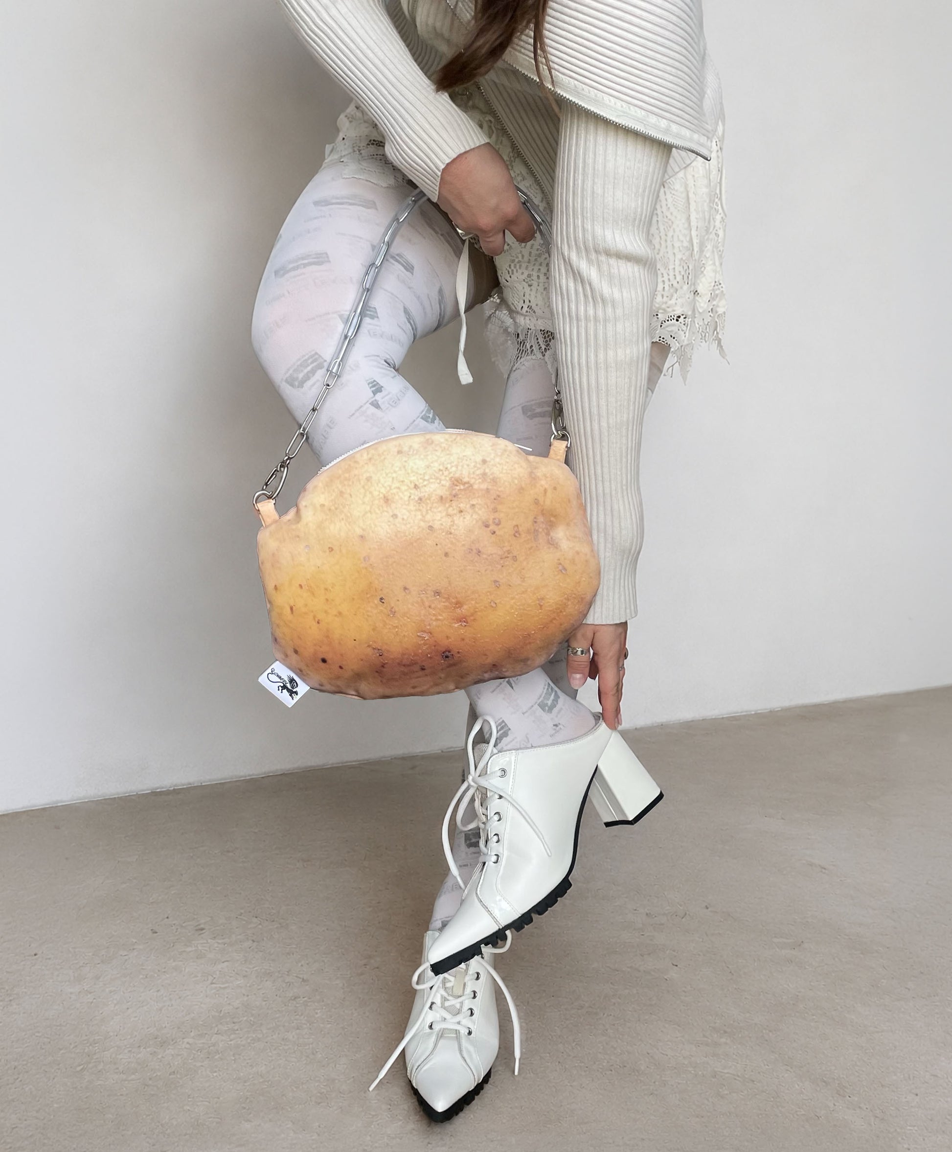 A person wearing a cream ribbed sweater, light patterned leggings, and white lace-up block-heel boots carries a large realistic potato-shaped shoulder bag against a plain light gray wall and concrete floor.
