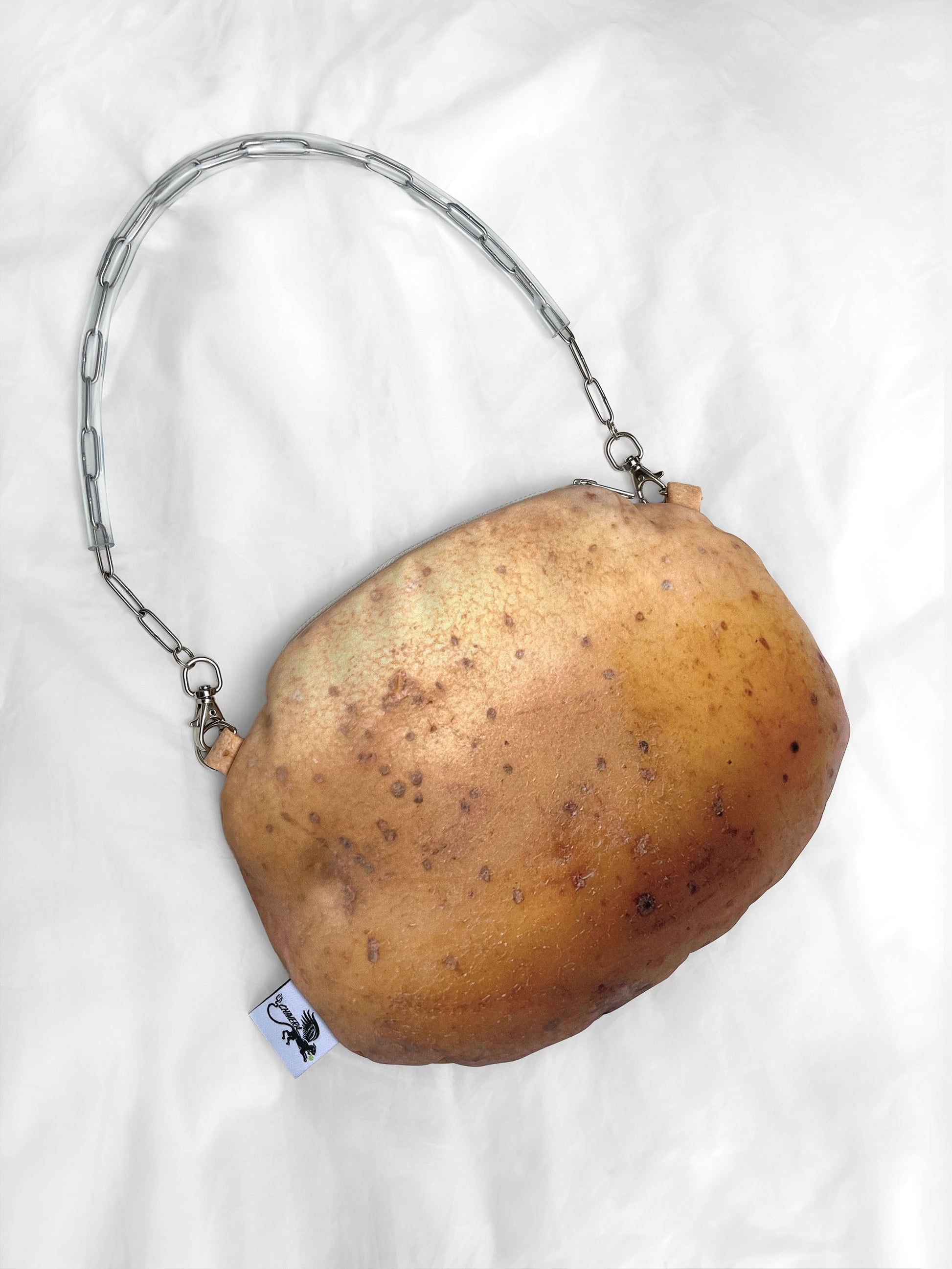 A novelty shoulder bag shaped like a potato, featuring a silver metal chain strap and a small white tag with a black logo, laid flat on a white fabric background.
