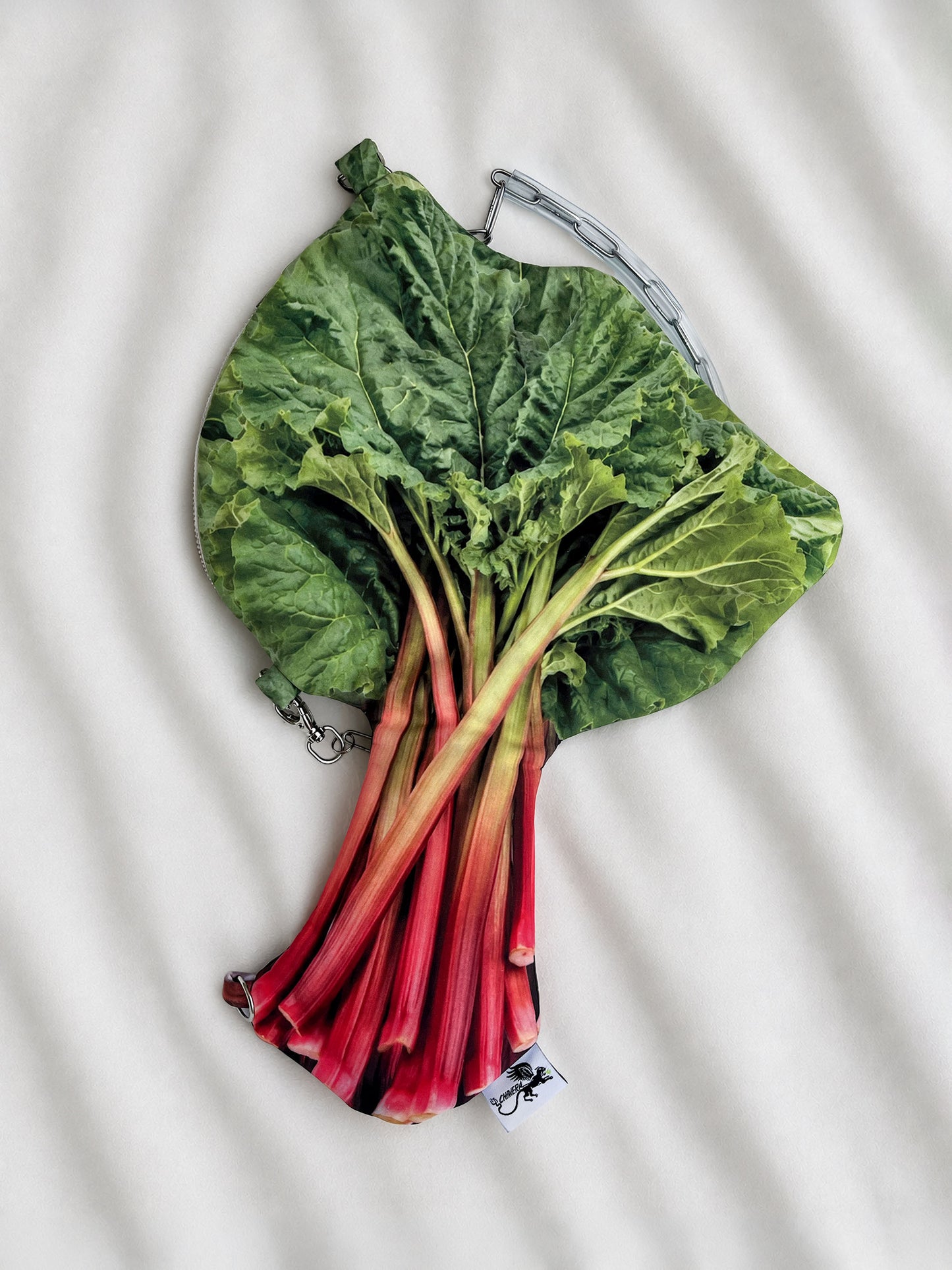 A novelty clutch purse designed to look like a fresh bunch of rhubarb, featuring green leaves and red stalks, laid flat on a white textured fabric background.