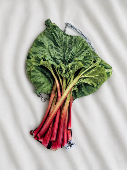 A novelty clutch purse designed to look like a fresh bunch of rhubarb, featuring green leaves and red stalks, laid flat on a white textured fabric background.