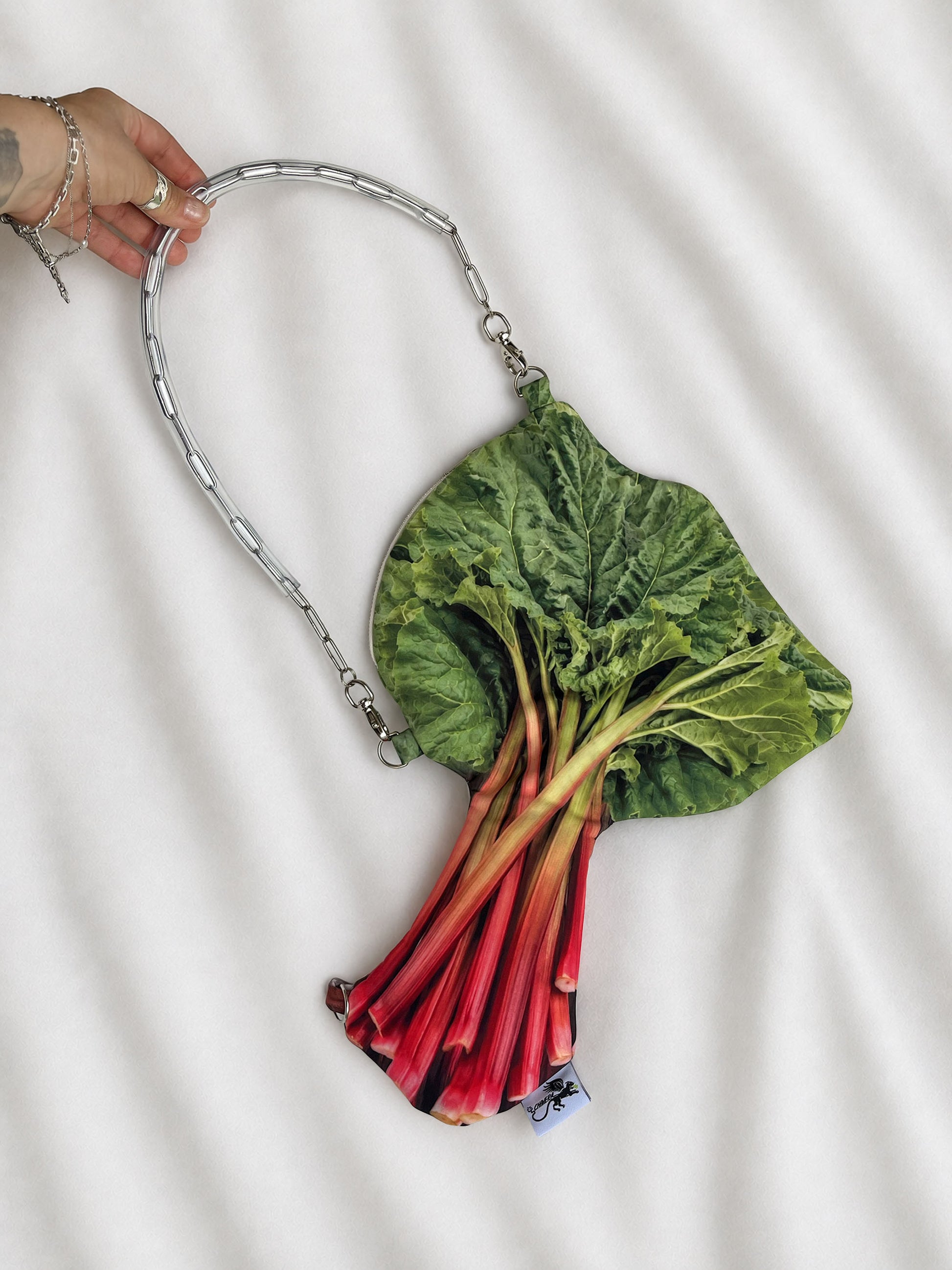 A hand holds a rhubarb-shaped shoulder bag with a silver chain strap against a white fabric background. The bag features a realistic print of green rhubarb leaves and red rhubarb stalks.