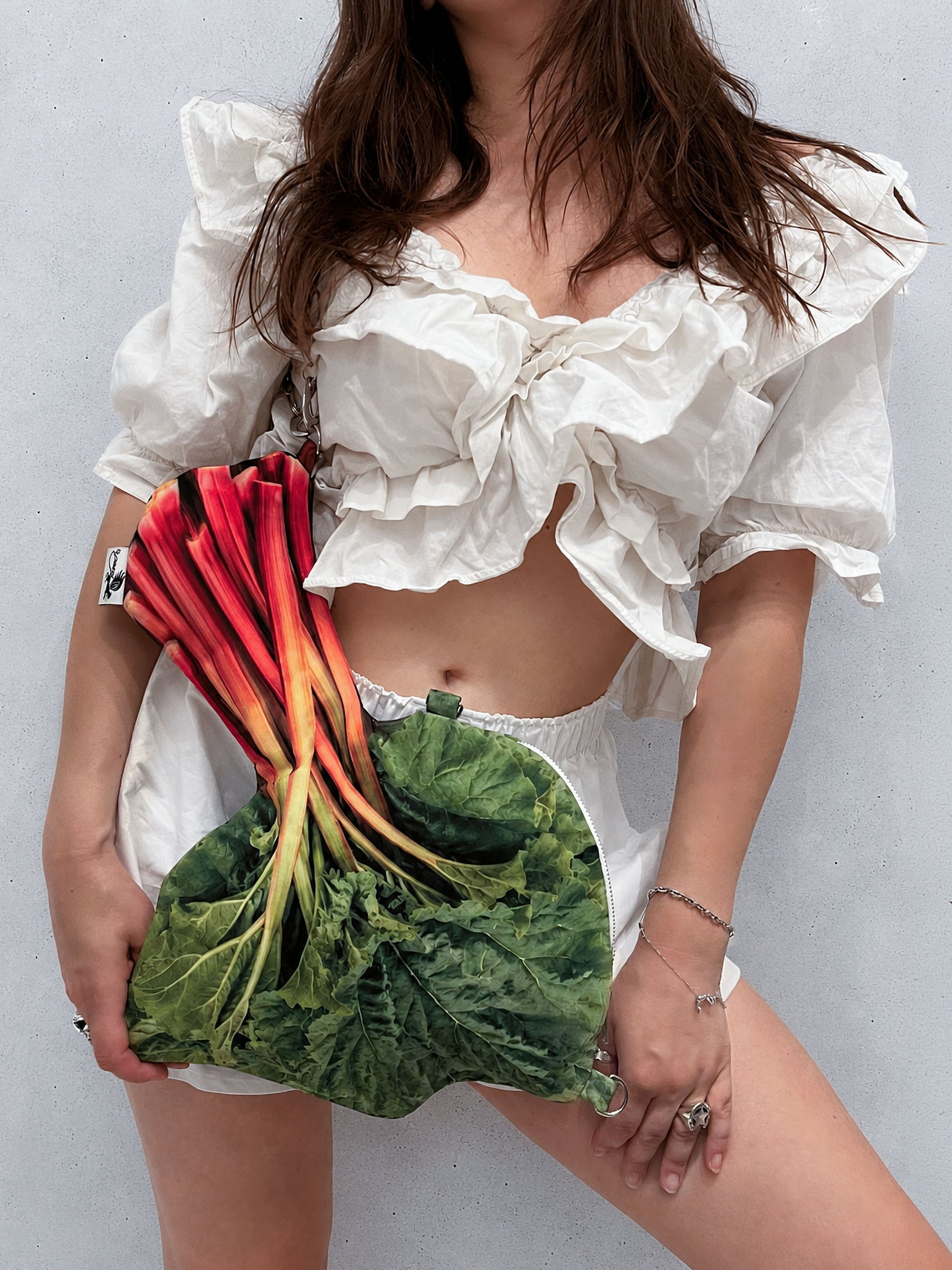 A model wearing a white ruffled crop top and white shorts, holding a novelty clutch bag shaped like a bunch of fresh rhubarb, set against a plain light gray wall.